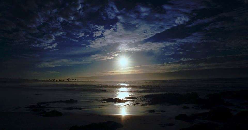 Here is image that is close to dark at Sunset. The sky has a dark ultramarine blue appearance. The clouds are hazy white, translucent with the dark blue sky. The upper part of the sky, you see a cumulus cloud whisping across the sky, and the lower clouds are hazy blending with the dark blue ocean below. The sun reflects on the ocean and the ocean has a dark teal blue appearance. It is a beautiful sunset.