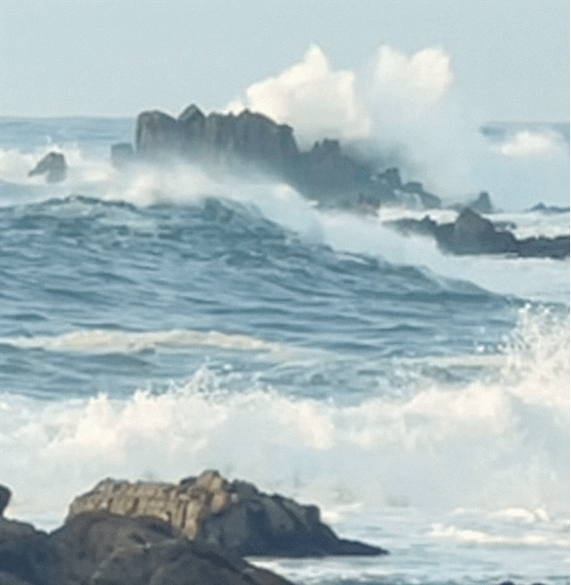 This gif image has moving ocean waves splashing high above a boulder in the middle of the ocean that's teal blue way out about 100 feet away from the rocky beach.