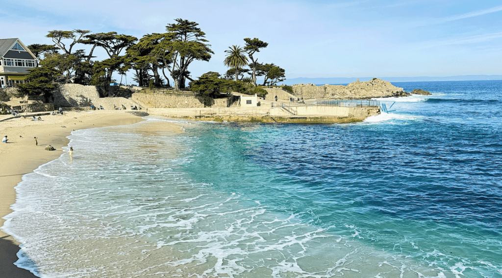 People relaxing on a clear day at Lover's Point.eautiful teal blue waves comes in to meet the light beige sand swishing with patterns on the misty white foam. There are cypress trees in the background at Lover's Point.