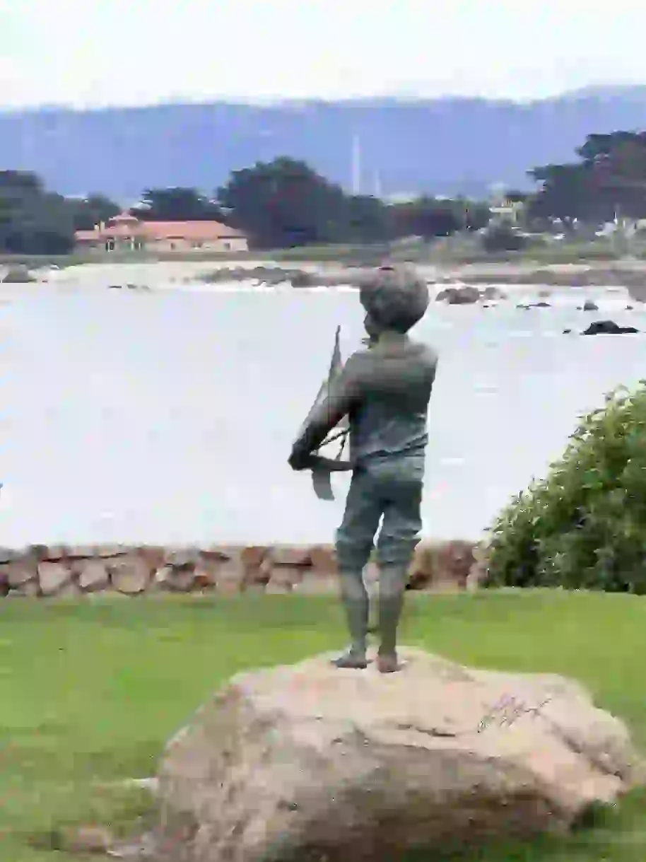 As far as the statue at Lover's Point, if you look In a distant, you can see the John Hopkins Marine Lab, behind that the aquarium, beyond that the Toro mountain range. A walking trail leads up to the viewers. The water is various shades of white and the sky is cloudy.