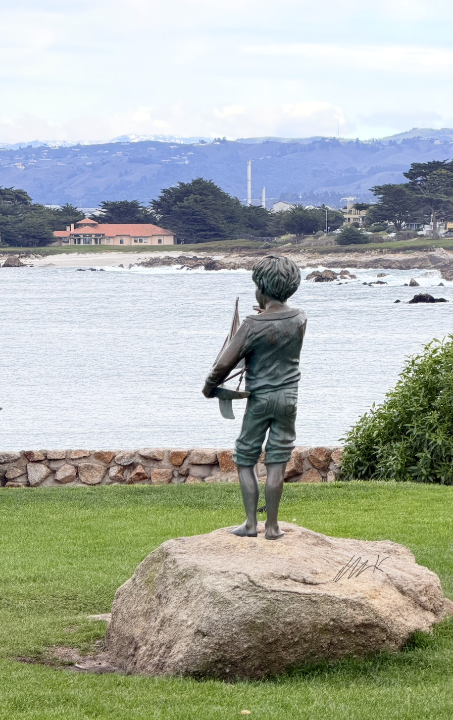 You see a scene at Monterey  Bay, at Lovers Point with a statue of boy, holding a sailboat both made out of bronze metal. The boy is staring towards the aquarium where you can see the mountains in the background with snow on top. The mountain is ultramarine light blue, there are Cypress trees along the beach and the water is a little hazy not yet showing some blue as it usually does. Thank you.