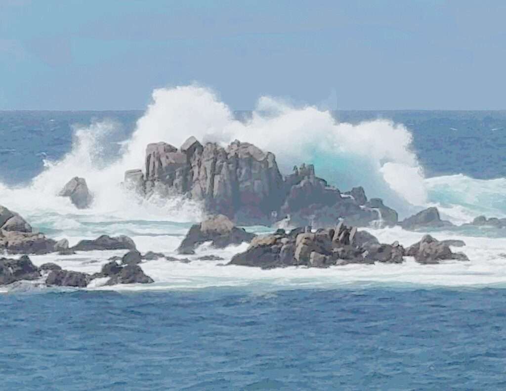 Out in the deep teal blue ocean is a  boulder, that is huge, where giant waves hit the boulder, causing so mud white foam and mist.