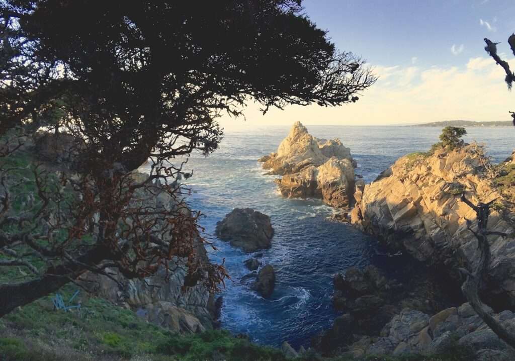 At the beach of Point Lobos State Park, with a windswept cypress tree, and rocks, that looks like a separate island separated by teal blue ocean water. There's partly cloudy skies.