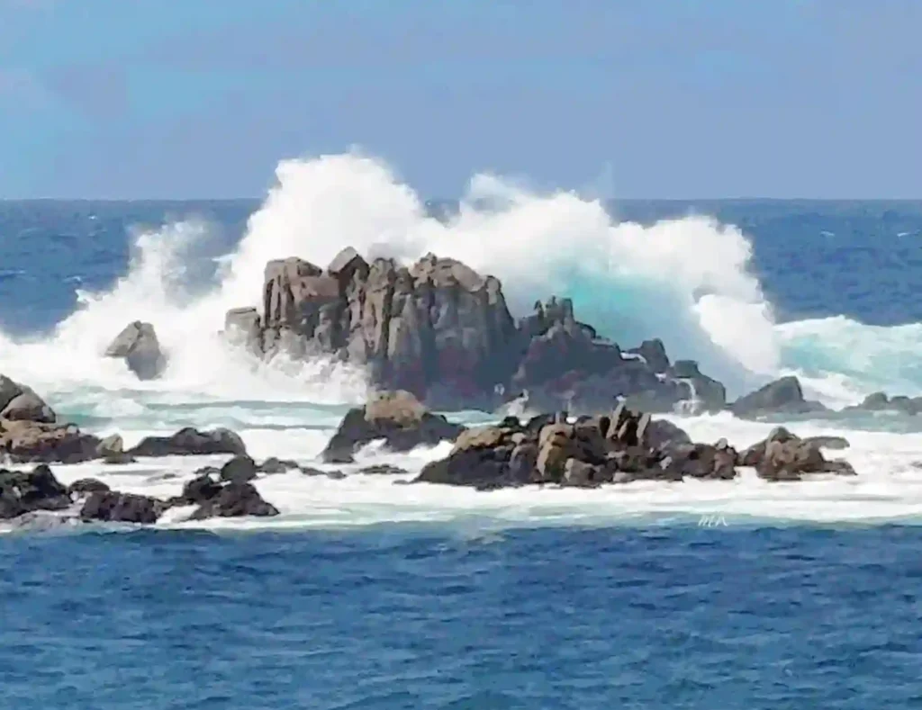 Out in the deep teal blue ocean is a  boulder, that is huge, where giant waves hit the boulder, causing so mud white foam and mist.