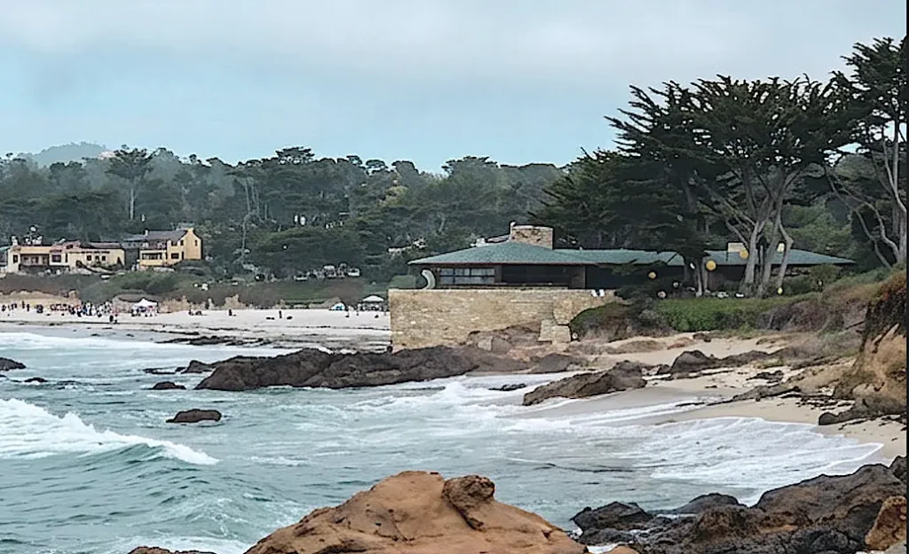 Walking along Carmel Beach, where the pristine, soft, white and powdery sand stretches out, there's a unique juxtaposition of homes perched on solid ground. Such as one resembling a ship’s bow cutting through the Pacific Ocean. It's built using local Carmel stone, with a green copper roof, it seamlessly blends with the teal blue sea and various burnt umber to reddish brown rocks, as if it is poised to embark on a journey. This architectural design, harmonize with their natural surroundings, appearing as if they have organically grown from the landscape. There are tall cypress trees surrounding the beach with light to medium grayish trunks and branched and hooker green pine needles and leaves. The sky is a beautiful medium blue with with lightly glowing translucent white closer to the top of the photo. There are some seaweed washed on the beach, burnt umber, with subtle dark green blends.