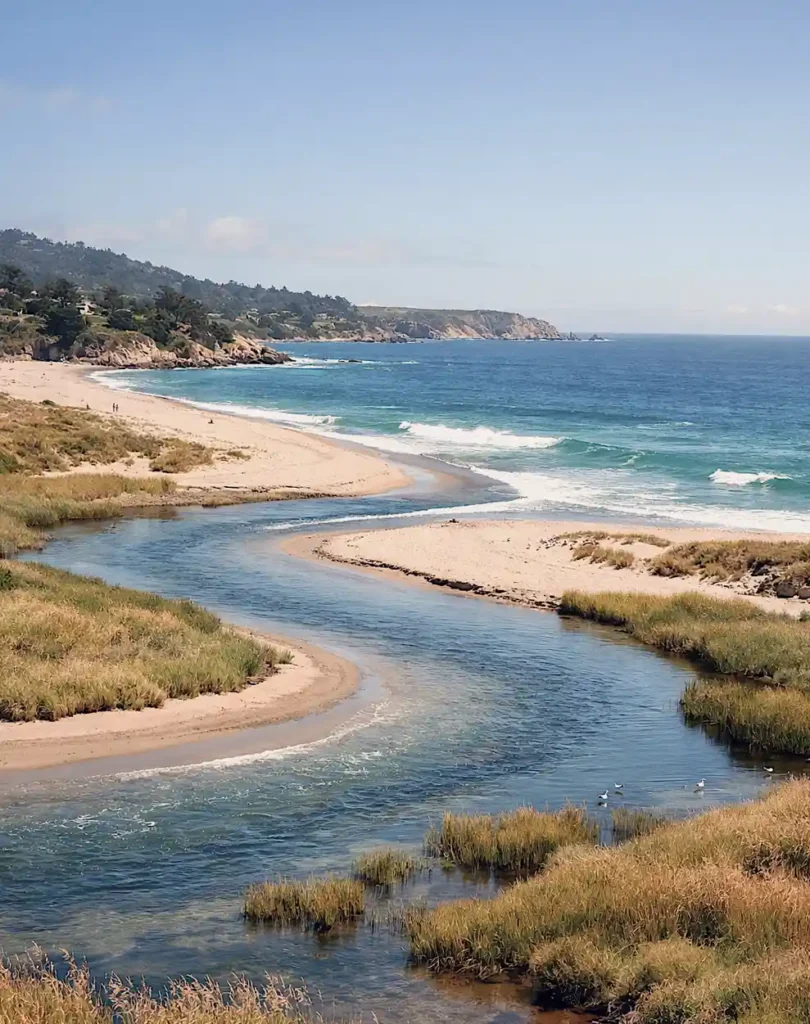 The landscape is defined by a deep bluish, light blue river emptying into the Carmel Bay with some rich teal blue at the ocean with highlights from the sky that is light blue. At the right the calm, fresh waters of the Carmel River begin to snake along the beach out to the ocean. Out toward the horizon, the powerful, crashing saltwater tides of the open Pacific carves with the fresh river water crashing through the sand that is light beige in color.
